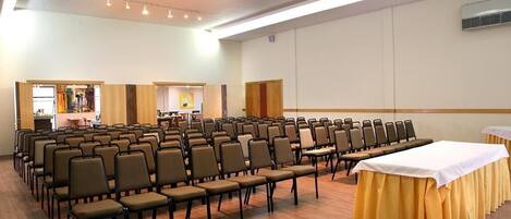 Un salón de conferencias con filas de sillas, una mesa larga con mantel blanco y un escenario con atril.