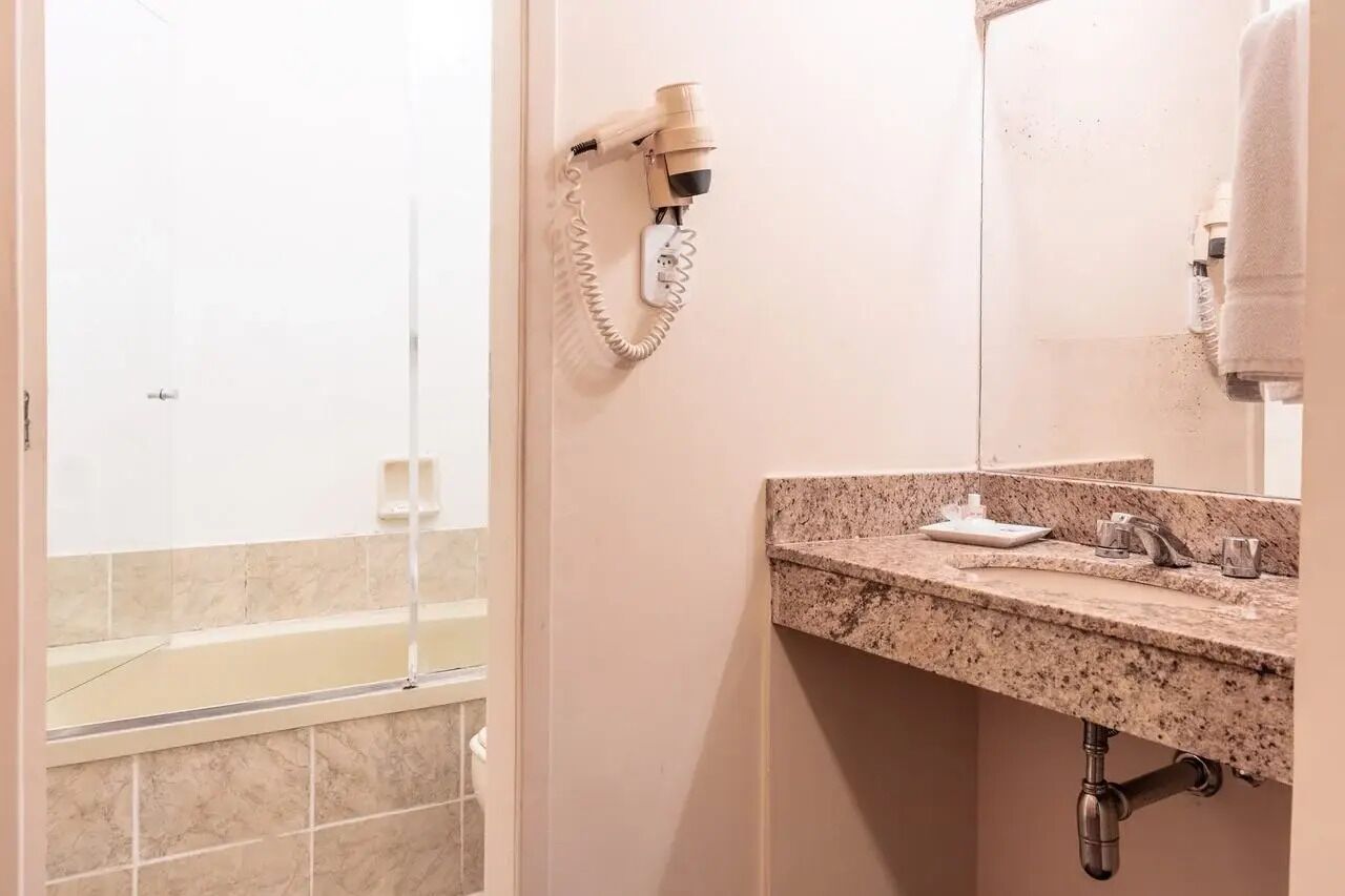 Un baño con lavamanos de mármol, un secador de pelo montado en la pared y una bañera visible a través de una puerta de cristal.