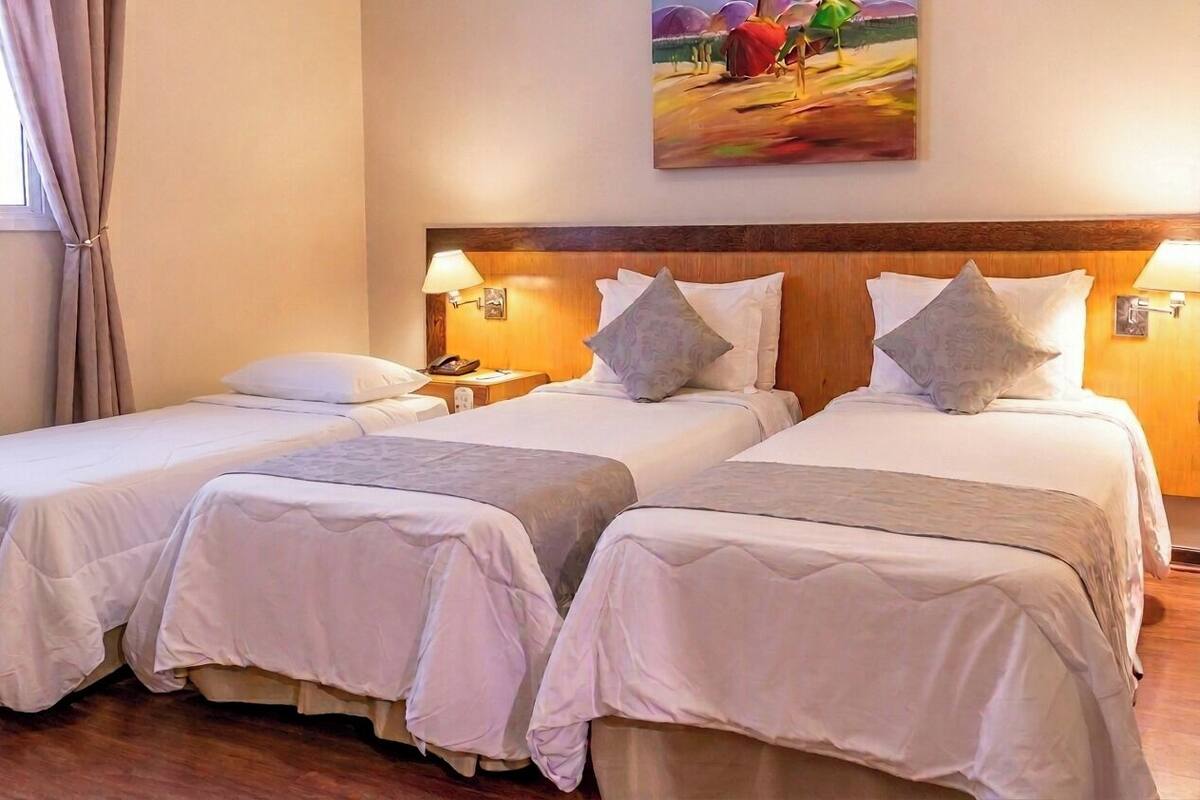 A hotel room with two single beds, a wooden headboard, bedside lamps, a painting on the wall, and a window with curtains.