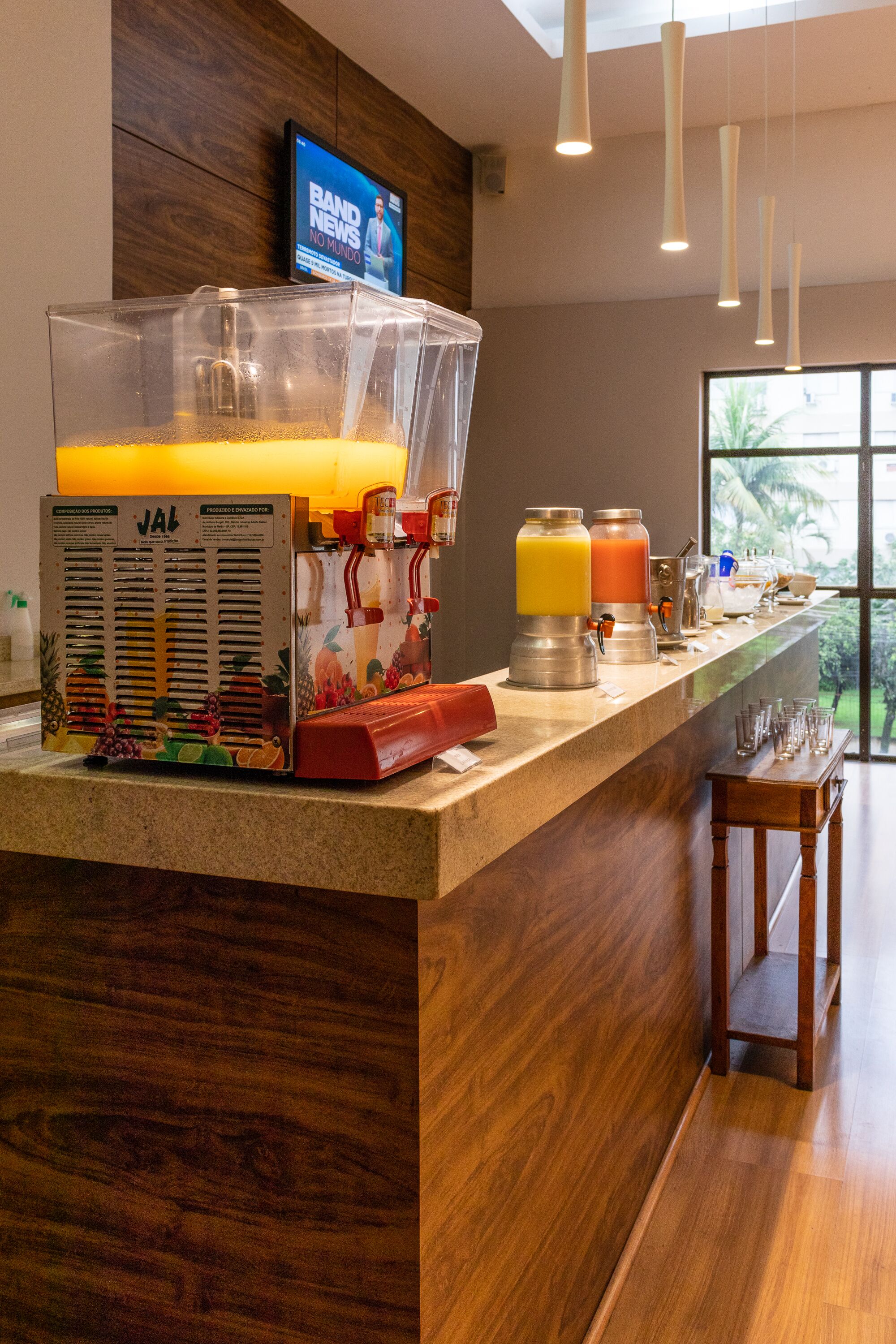 A breakfast bar with a fruit press and juice dispensers, a television displaying news, and a view of the outdoors.