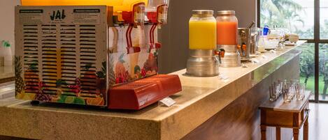 A breakfast bar with a fruit press and juice dispensers, a television displaying news, and a view of the outdoors.