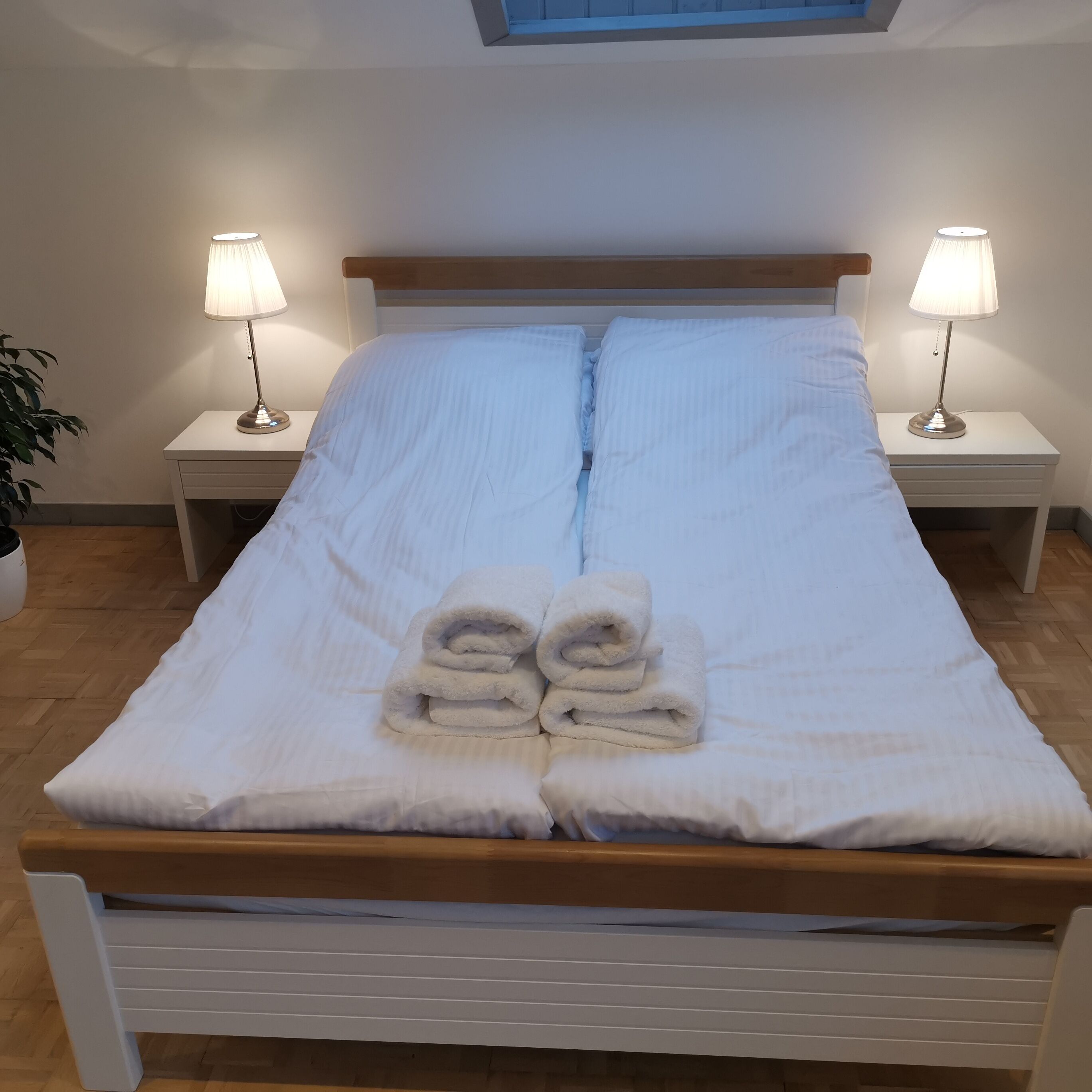 A neatly made bed with white linens and rolled towels, flanked by two bedside lamps on matching nightstands.