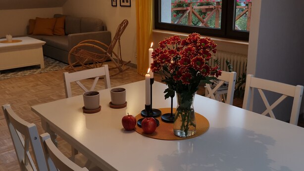 Uma área de refeições com uma mesa posta para dois, um vaso de flores e vista para uma paisagem montanhosa através de uma janela.
