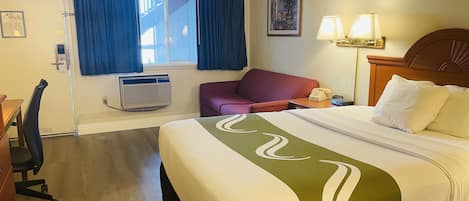 A hotel room with a bed, a desk, a chair, a window with curtains, a wall-mounted lamp, and a framed picture.
