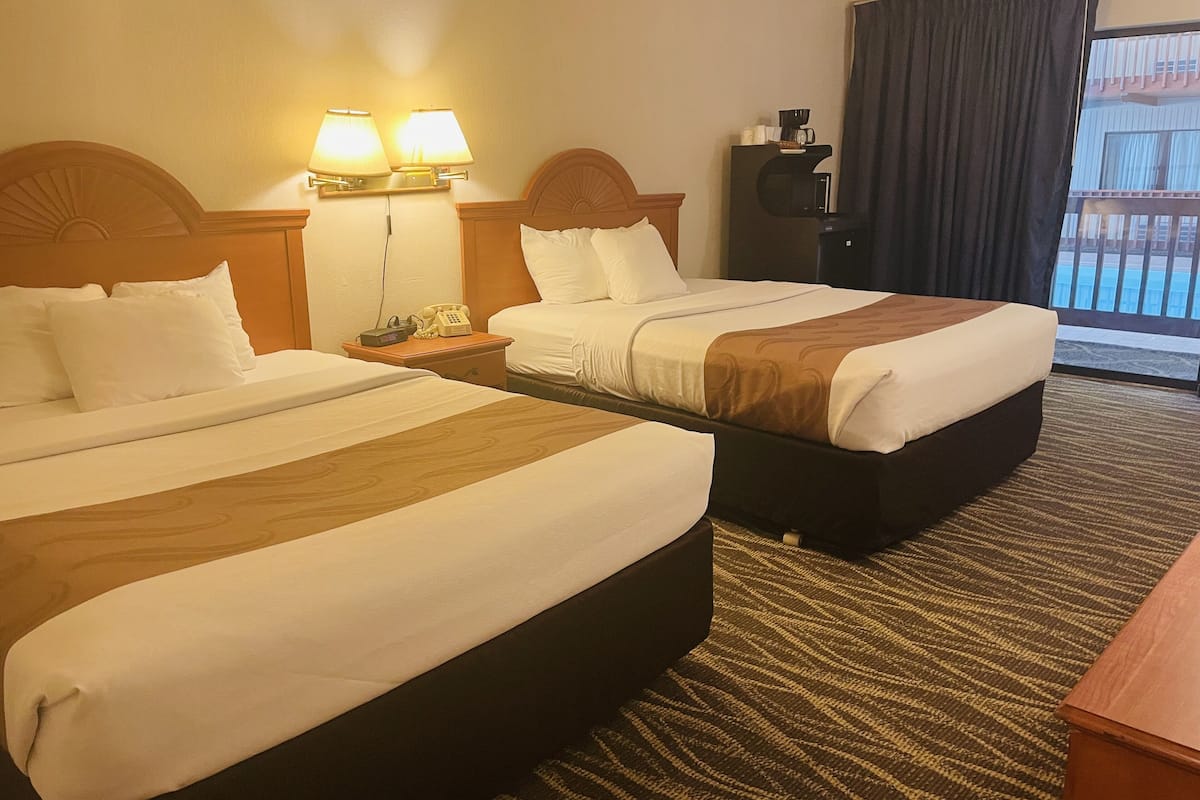 A hotel room with two beds, a nightstand with a lamp, a coffee maker, and a view of the outside balcony.