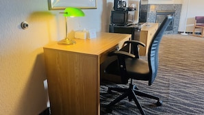 Habitación de hotel con un escritorio de madera, una silla de oficina negra, una lámpara de escritorio verde, una cafetera y un televisor sobre un soporte.