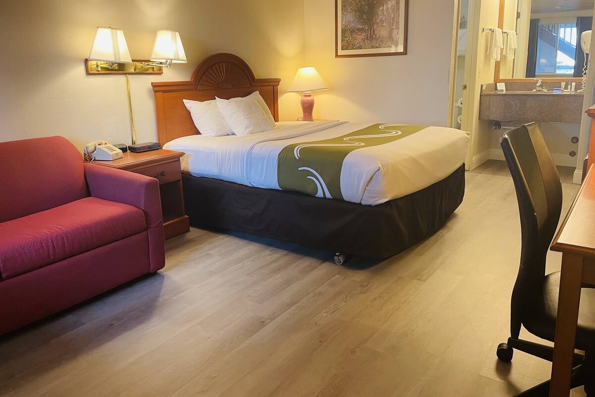 A hotel room with a bed, a red armchair, a desk, a chair, a nightstand, a lamp, a painting, and a bathroom with a mirror and towel rack.