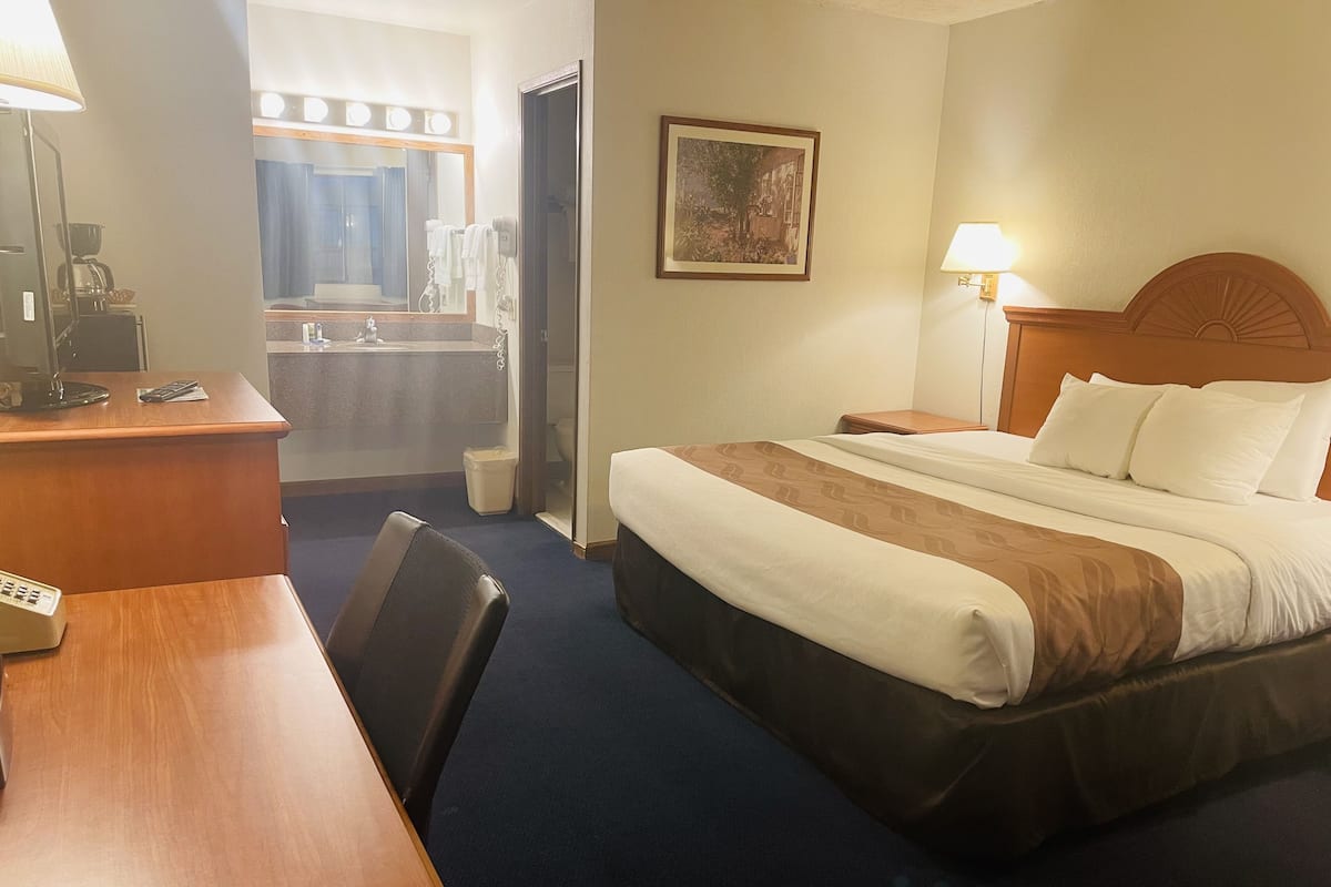 A hotel room with a bed, a desk with a lamp, a chair, a bathroom with a mirror, and a picture on the wall.