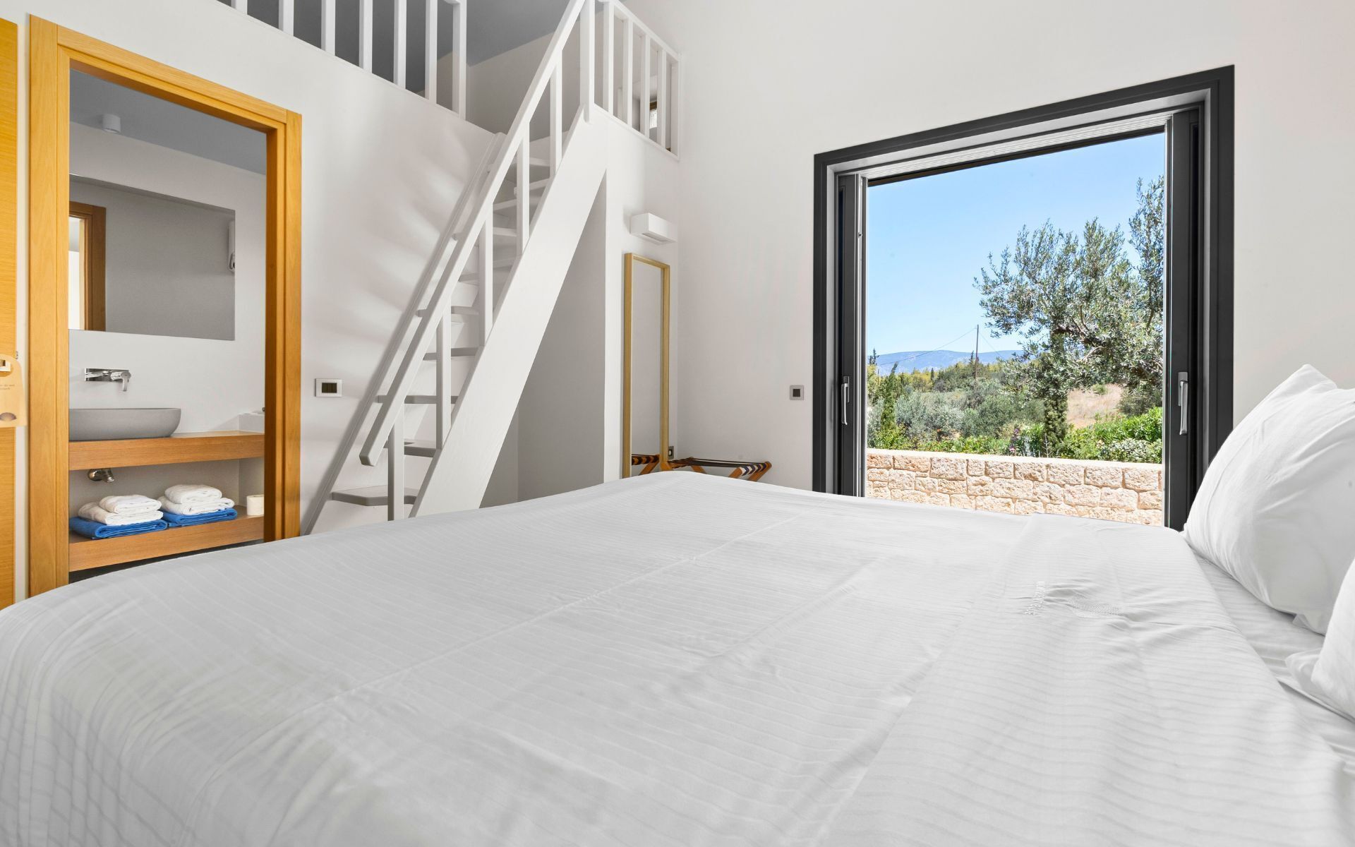 A bedroom with a bed, a bathroom with a sink and towel rack, a staircase, and a view of the outdoors.
