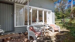 A covered porch with wicker chairs and a table, a grey door, and a window with curtains. - Crabapple Southside Mountain Cottage Pet Friendly (WENTWORTH FALLS)
