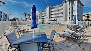 A poolside area with lounge chairs and umbrellas, a pool, and a multi-story building in the background. - Royal Pelican 173 by Distinctive Beach Rentals (Fort Myers Beach)
