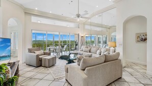 Un salón amplio con un sofá grande, una mesa de centro y un televisor. Hay un balcón con ventanas grandes y vistas al exterior.