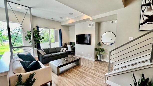 Een moderne woonkamer met een bank, salontafel en een televisie. Er is een trap met een leuning, en er staan potplanten.