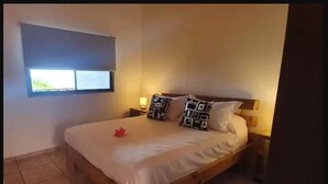 A single bed with a white mattress and patterned pillows, a nightstand with a lamp, and a window with a view of greenery. - Tupuna Lodge Apartments (Hanga Roa)