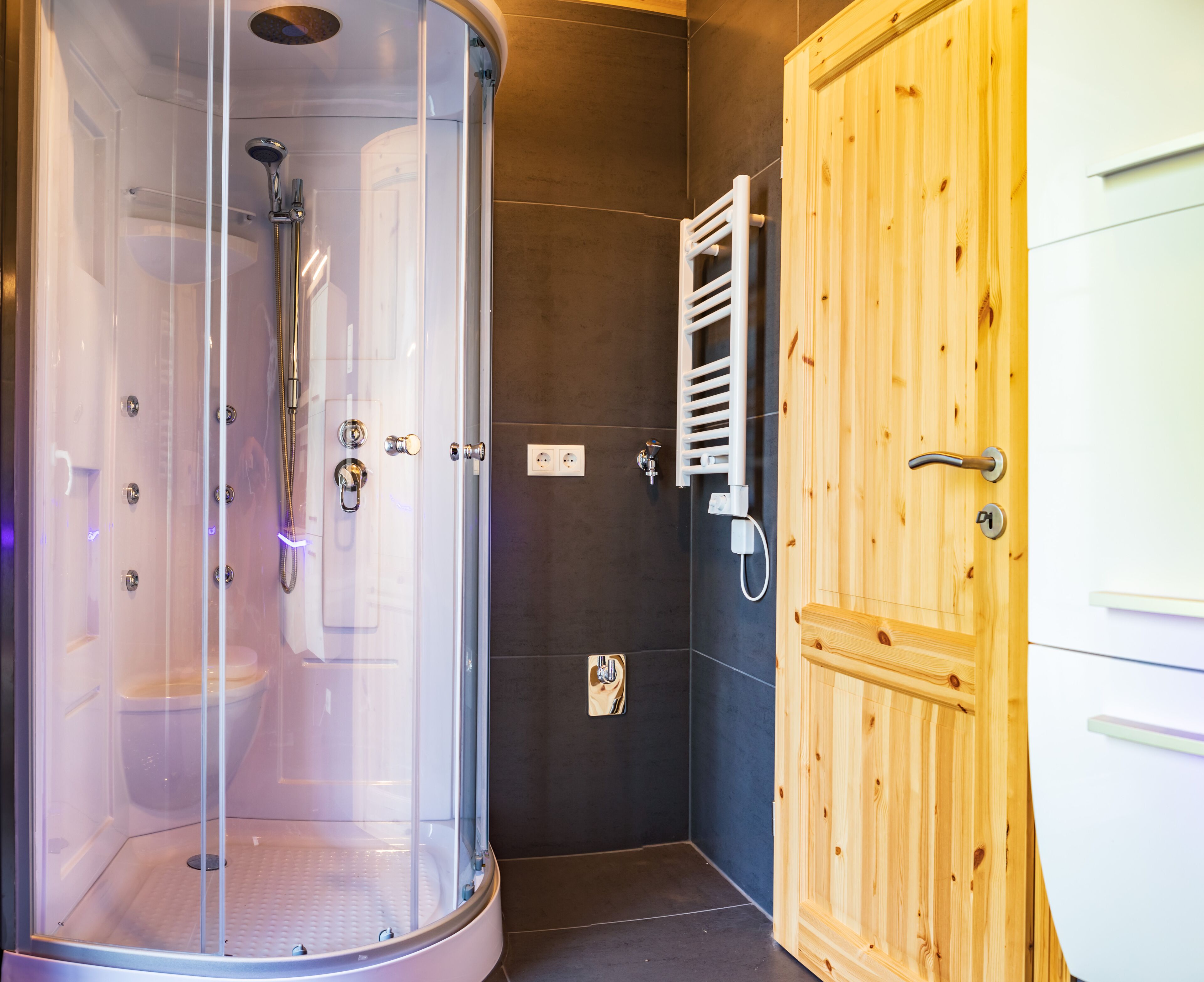 A shower enclosure with a glass door, a wooden door with a towel rack, and a white cabinet.