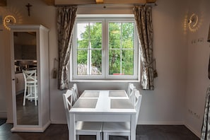A dining area with a white table and chairs, a window with curtains, and a view of greenery outside.