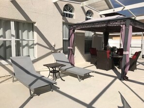 A covered outdoor seating area with lounge chairs and a table, a pergola with curtains, and a building with large windows in the background. - 4BR Davenport Pool Home by Watson-727RL (Davenport)