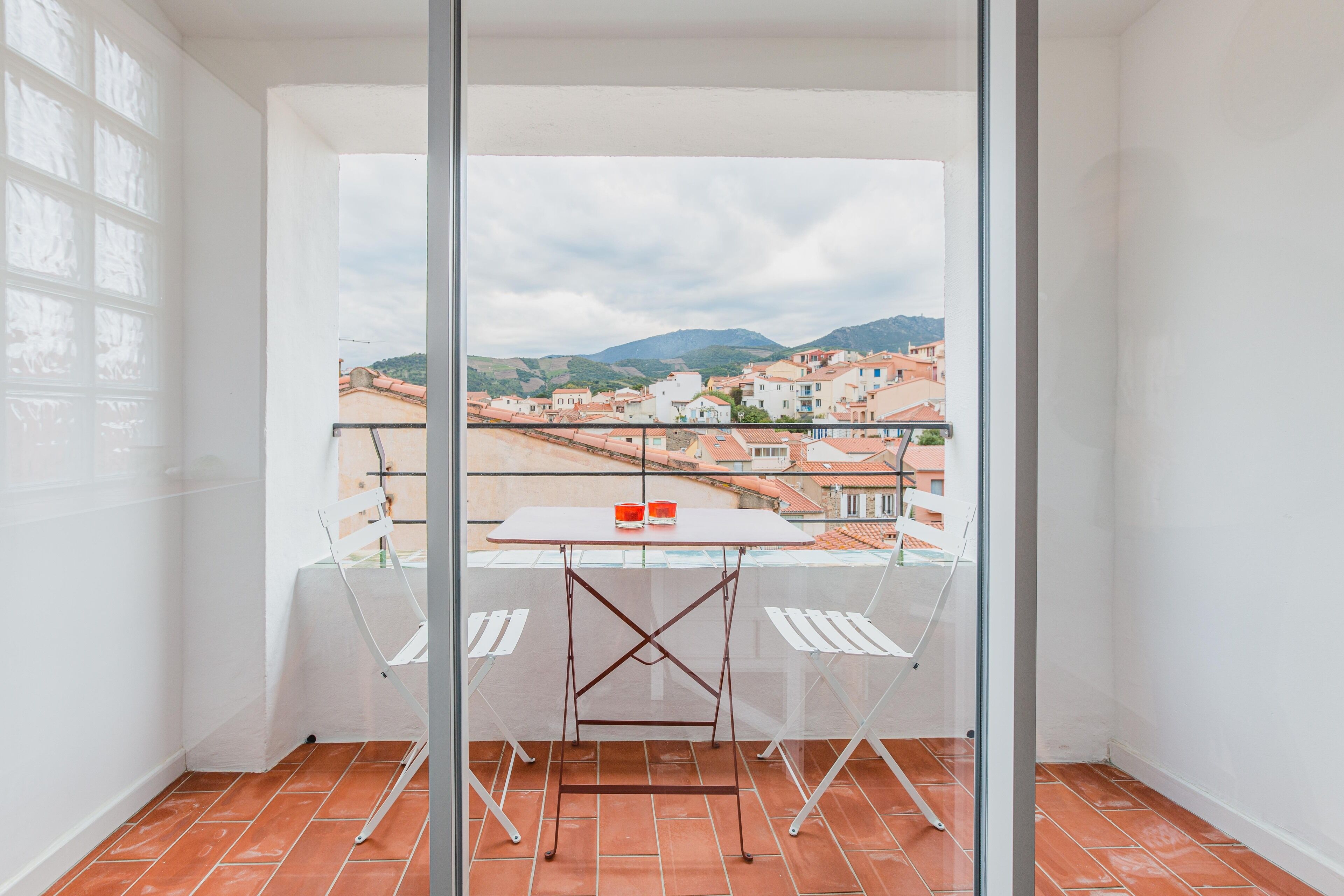 A balcony with a table and chairs, overlooking a town with red-roofed buildings and hills in the distance.
