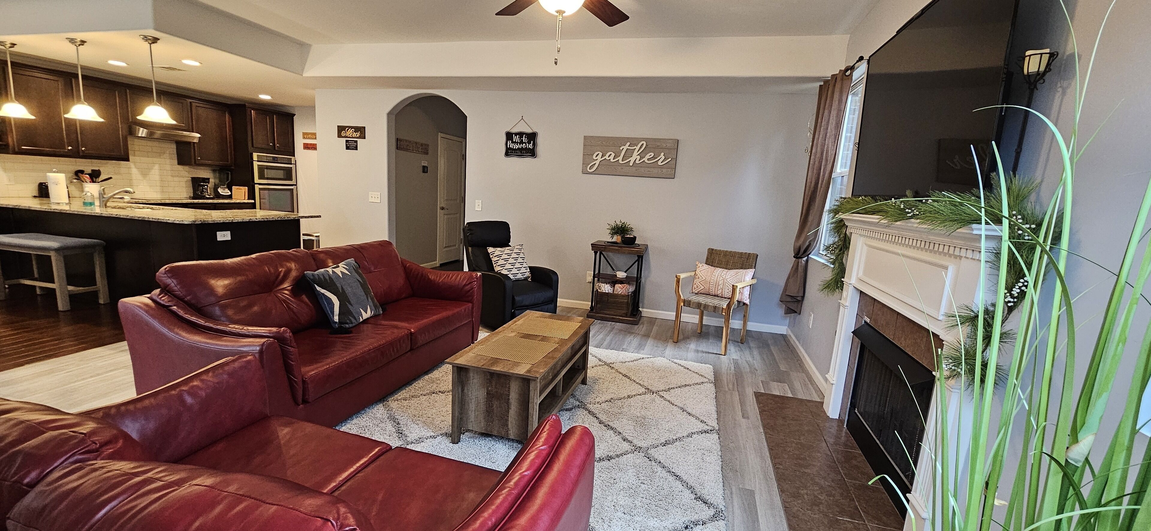 A modern living room with a red leather sofa, a wooden coffee table, and a fireplace. There is a kitchen in the background with dark wooden cabinets and stainless steel appliances.