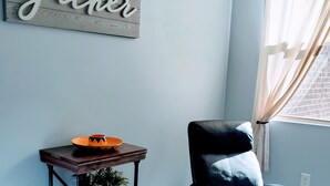 A room with a dark blue armchair, a wooden side table with a bowl and a potted plant, and a wall sign that reads "gather".