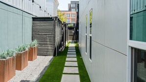 Une ruelle étroite avec un chemin bordé de gazon synthétique vert, des jardinières en bois et un portail en métal.