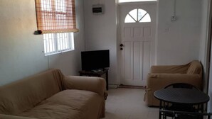 Une pièce avec un canapé beige, une petite table et une chaise, un téléviseur et une porte blanche avec une fenêtre.