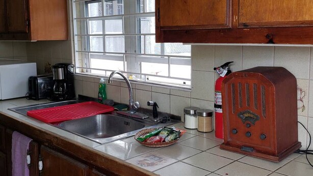 Une cuisine avec des meubles de rangement en bois, une fenêtre, un évier, une machine à café, une radio et un tapis rouge.