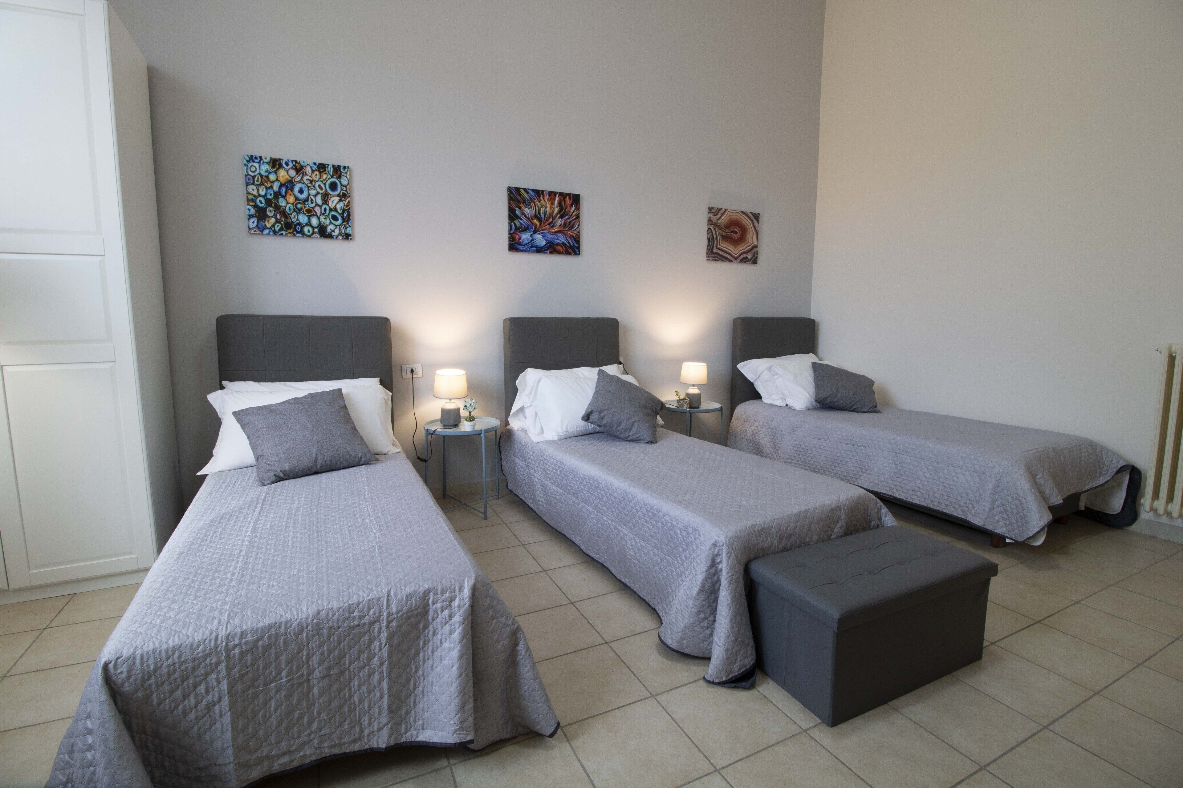 Ein Hotelzimmer mit drei Einzelbetten, jedes mit grauer Bettdecke und Kissen. An den Nachttischen stehen Lampen und darüber befinden sich Wandbilder.