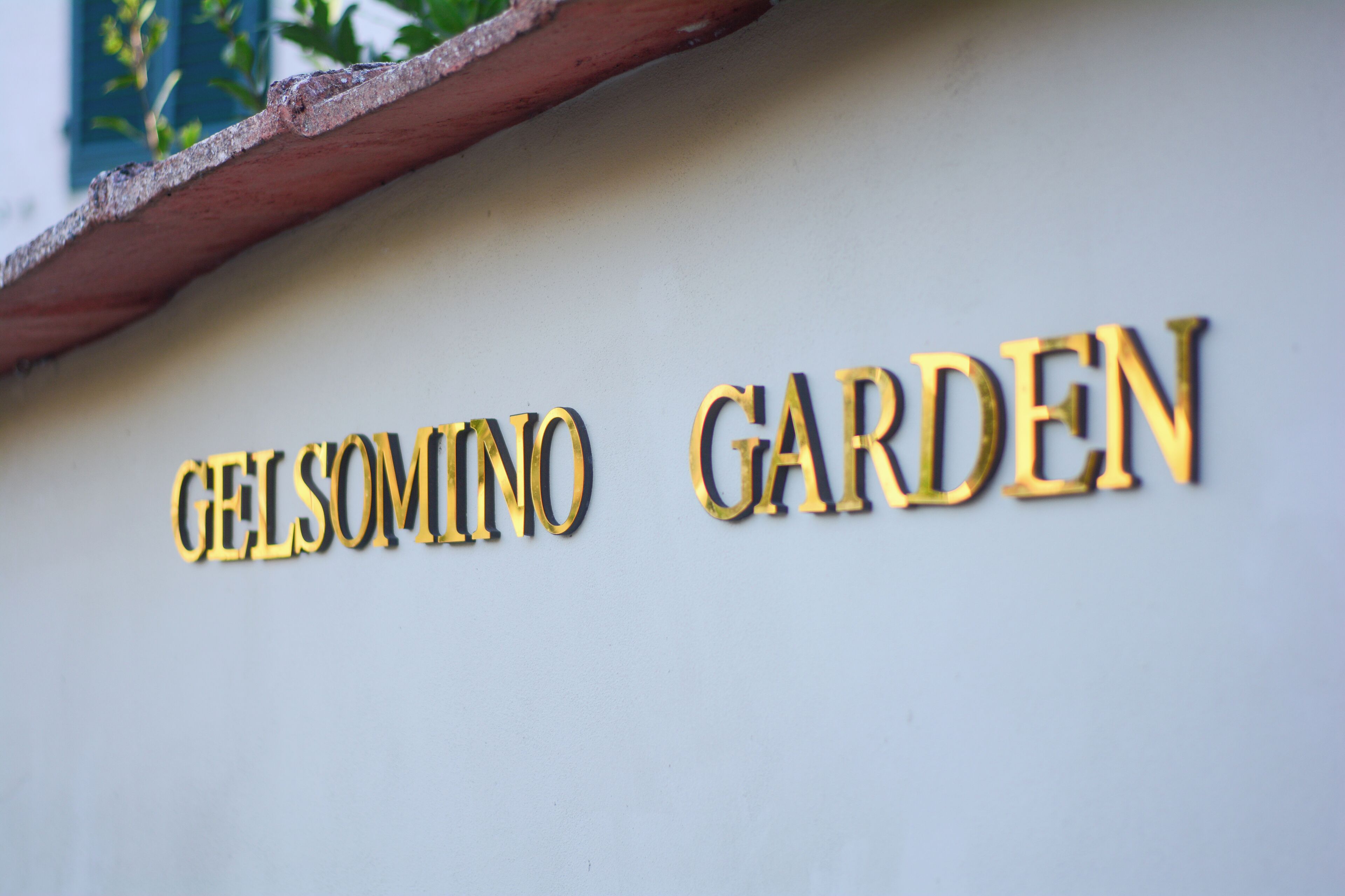 A sign with the words "GELSOMINO GARDEN" in gold lettering on a white background.