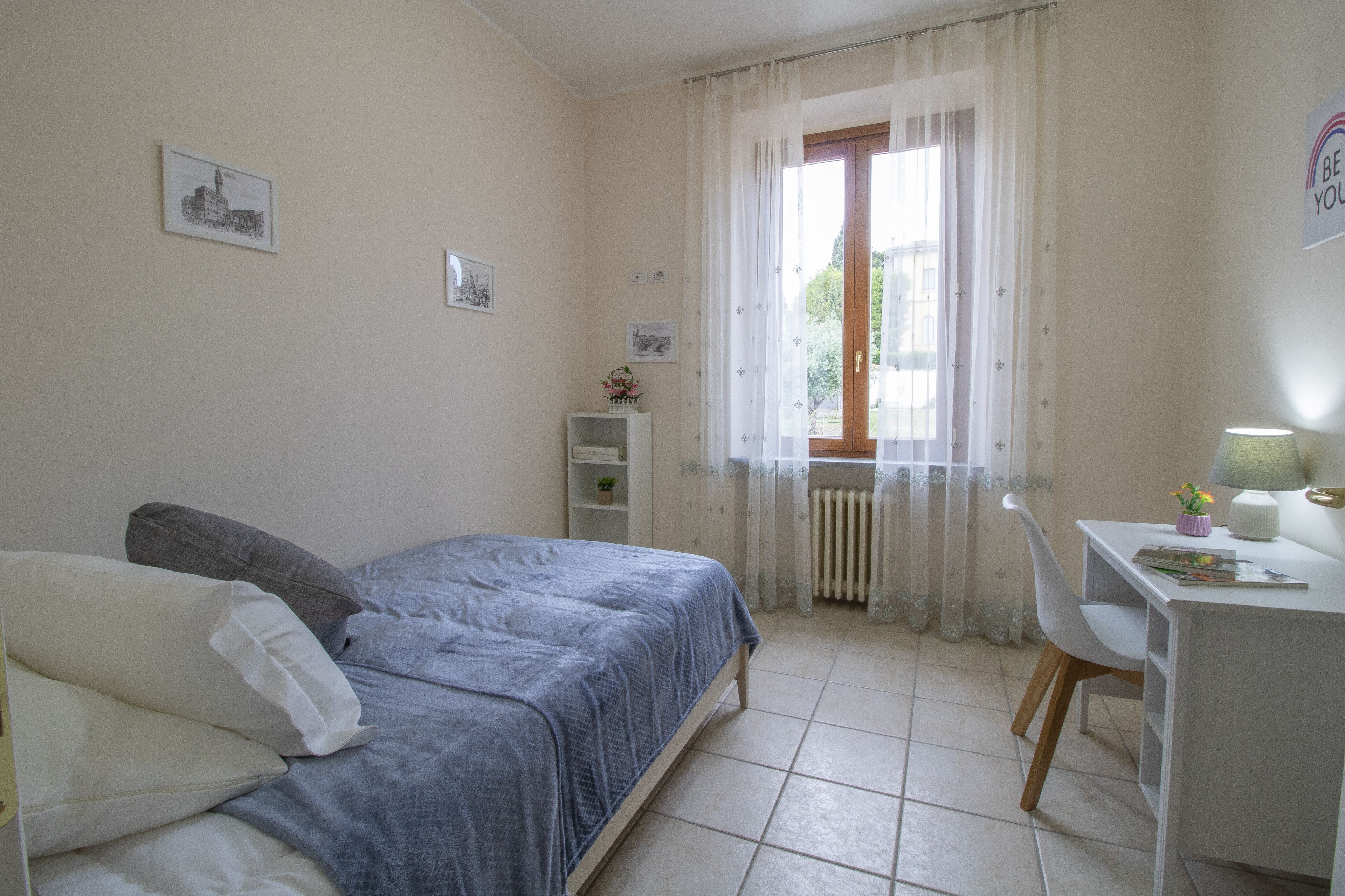 Une chambre à coucher avec un lit, un bureau et une chaise. Il y a des rideaux à la fenêtre, et on peut voir un radiateur.