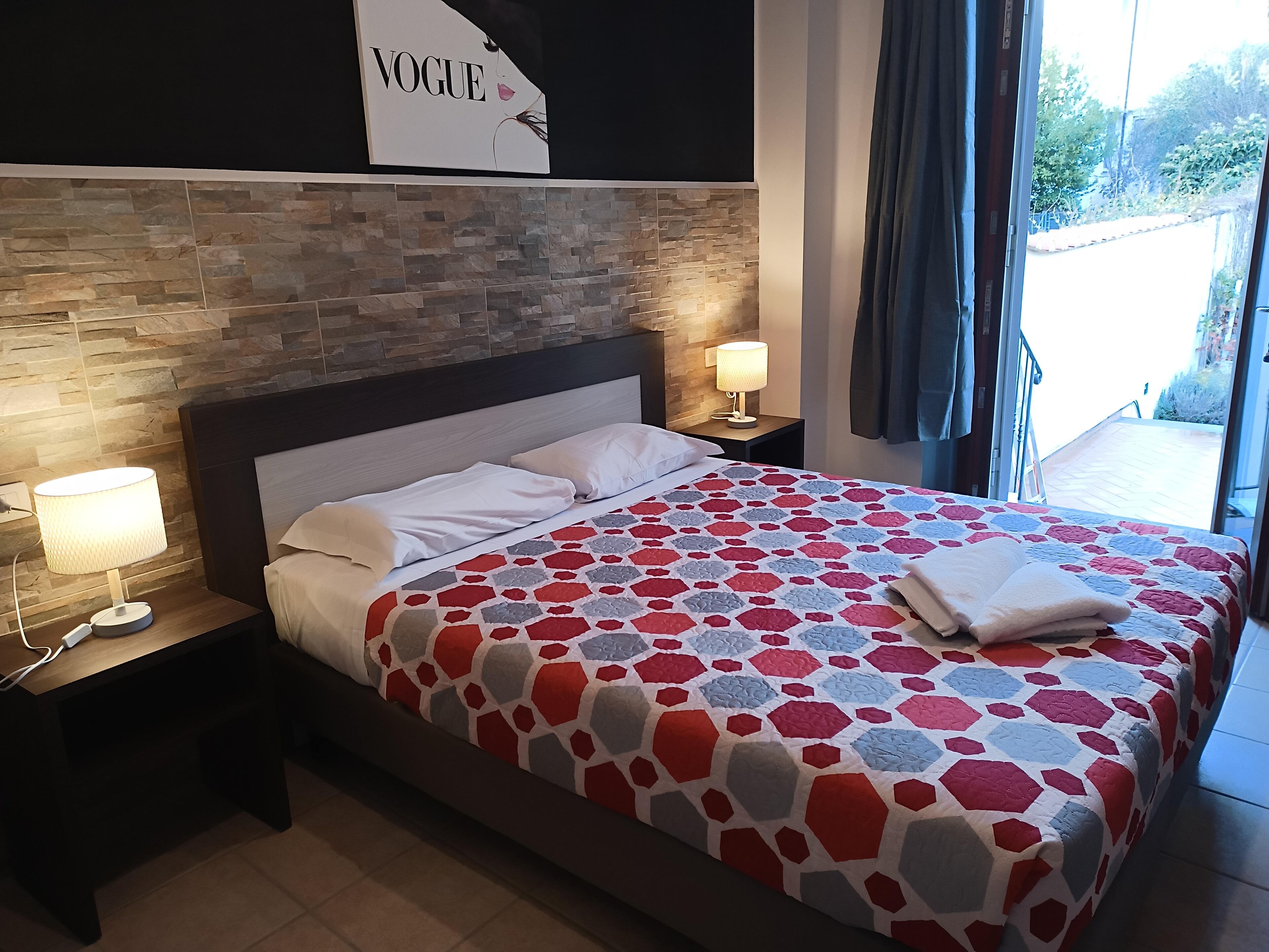 Een hotelkamer met een muur in steenmotief, een bed met een rood- en grijspotten dekbed, twee bedlampjes en een 'VOGUE' bord op de muur.