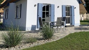 Una casa blanca con contraventanas azules, techo de paja y chimenea. Cuenta con un patio con dos sillas y una mesa.
