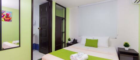 Habitación de hotel con cama, mesita de noche con teléfono y planta, espejo y pared verde.