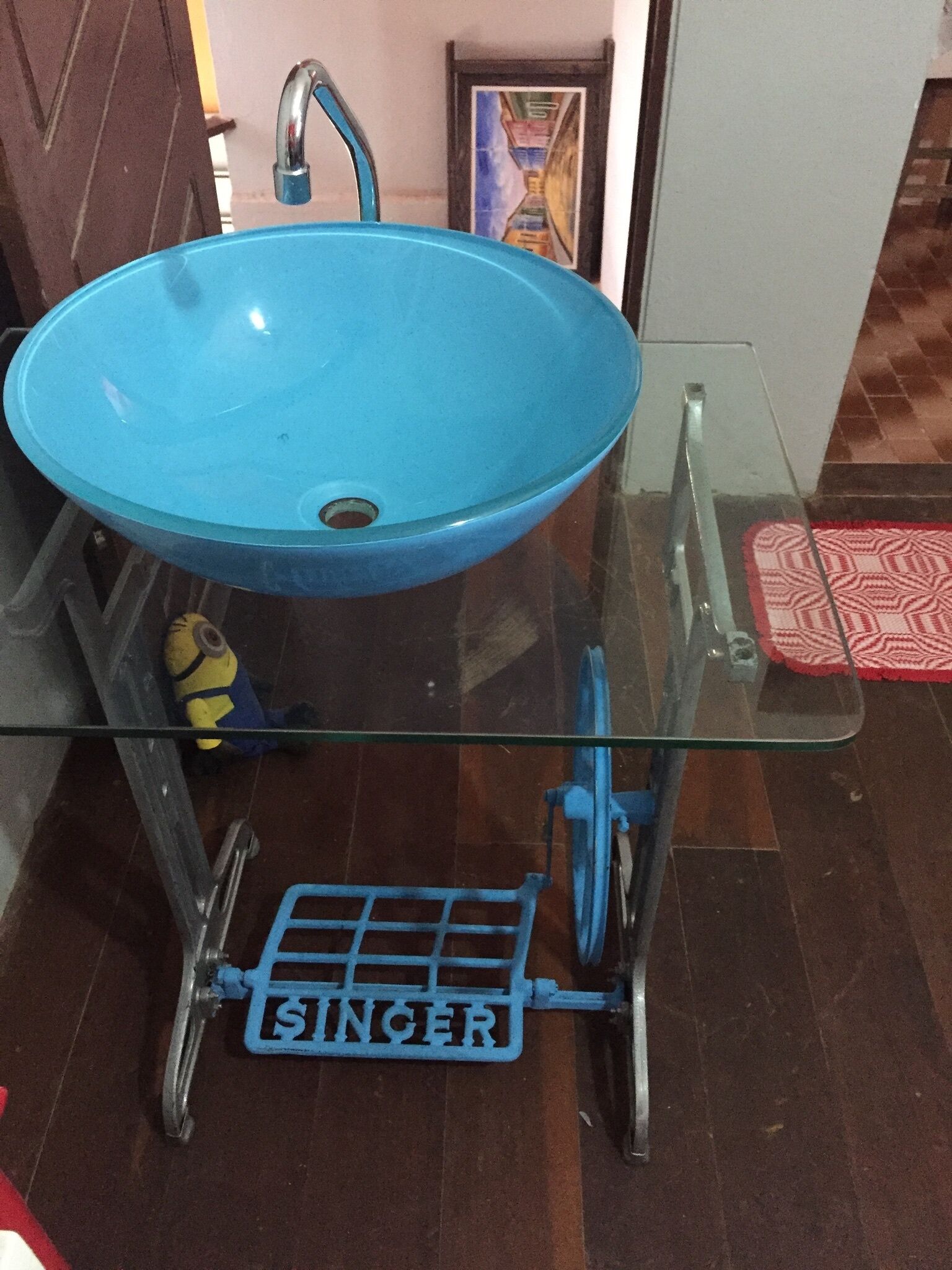 Un baño con un lavabo azul sobre un estante de vidrio, un taburete metálico con la palabra "SINGER" y un felpudo rojo con estampado en el suelo.