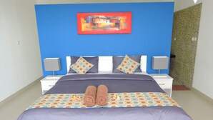 A neatly made bed with a patterned bedspread and two rolled towels, a blue wall with a colorful abstract painting, and a white nightstand with a lamp. - Kartika Villas (Buleleng)