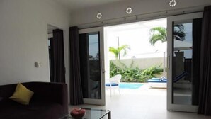A living room with a dark-colored sofa, a glass coffee table, and a sliding glass door leading to a pool area. - Kartika Villas (Buleleng)