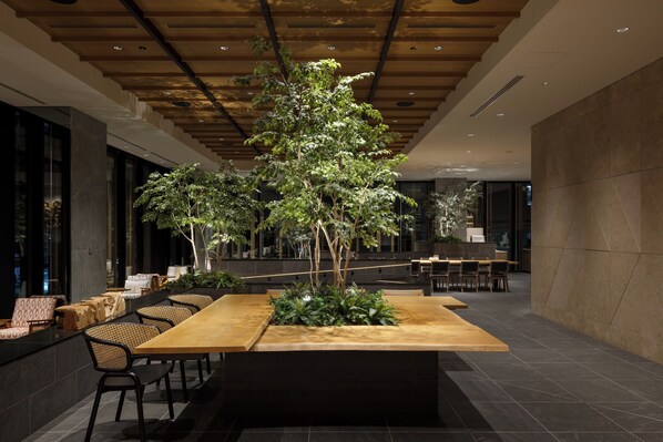 A modern indoor space with a long wooden table surrounded by chairs, a central plant, and a seating area with cushioned chairs.