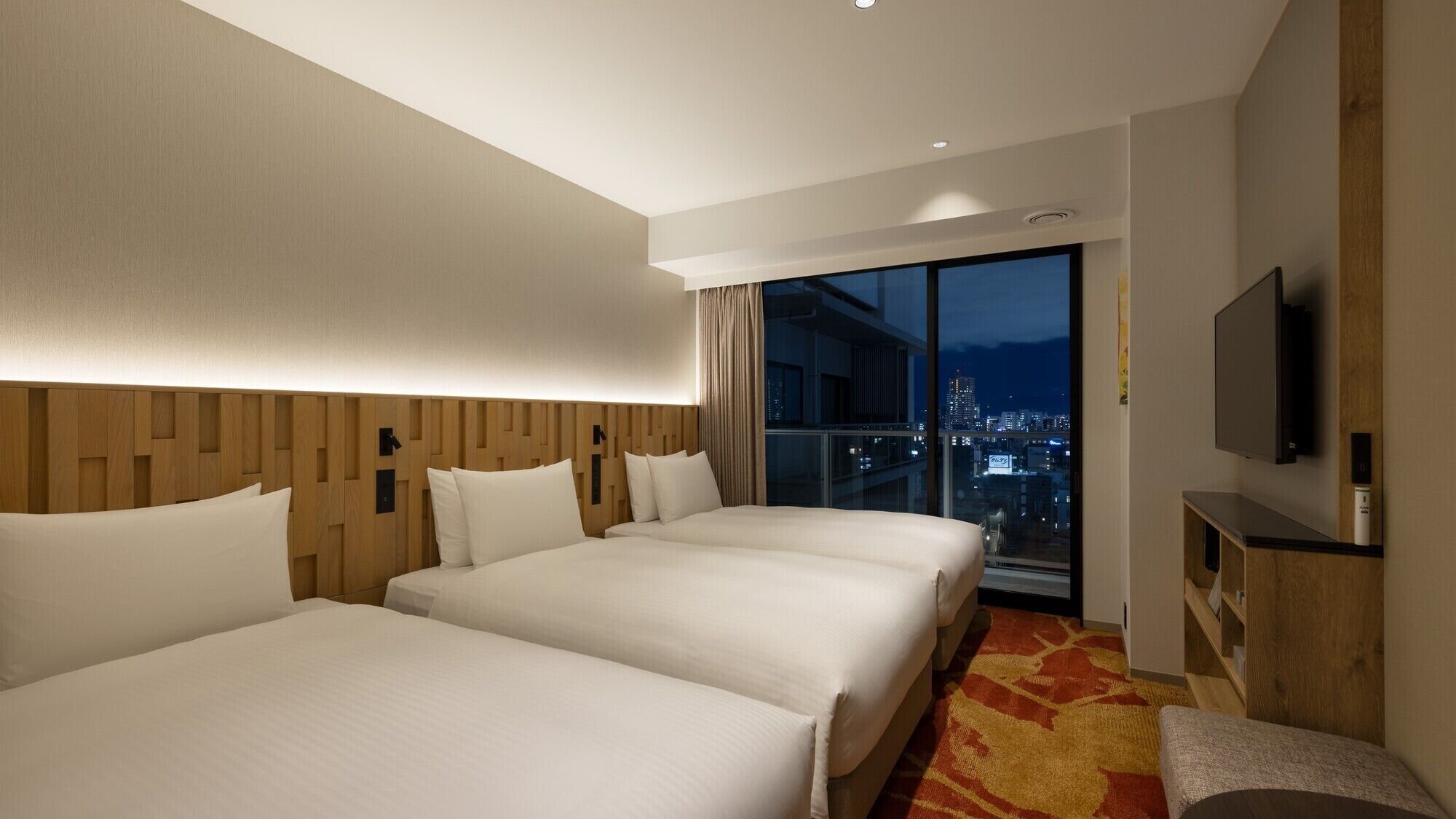 A hotel room with two beds, a TV, a large window with a city view, and a wooden headboard.
