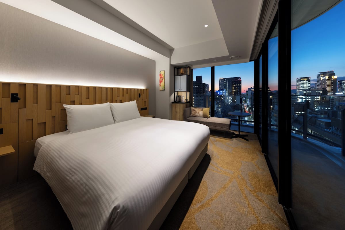 Een moderne hotelkamer met een groot bed, een zithoek en uitzicht op de stad door grote ramen.
