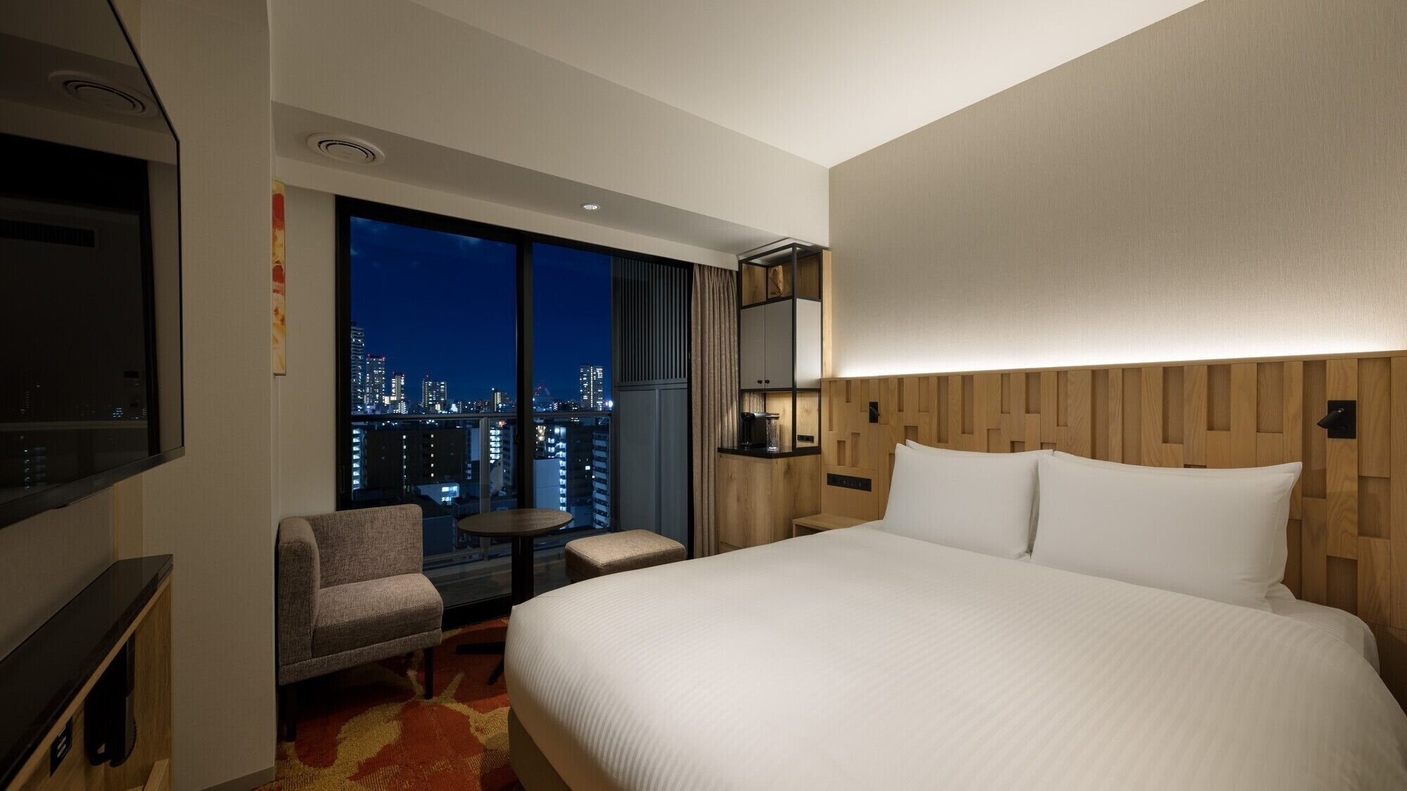 A hotel room with a large bed, a TV, a small table, a chair, and a view of the cityscape at night.