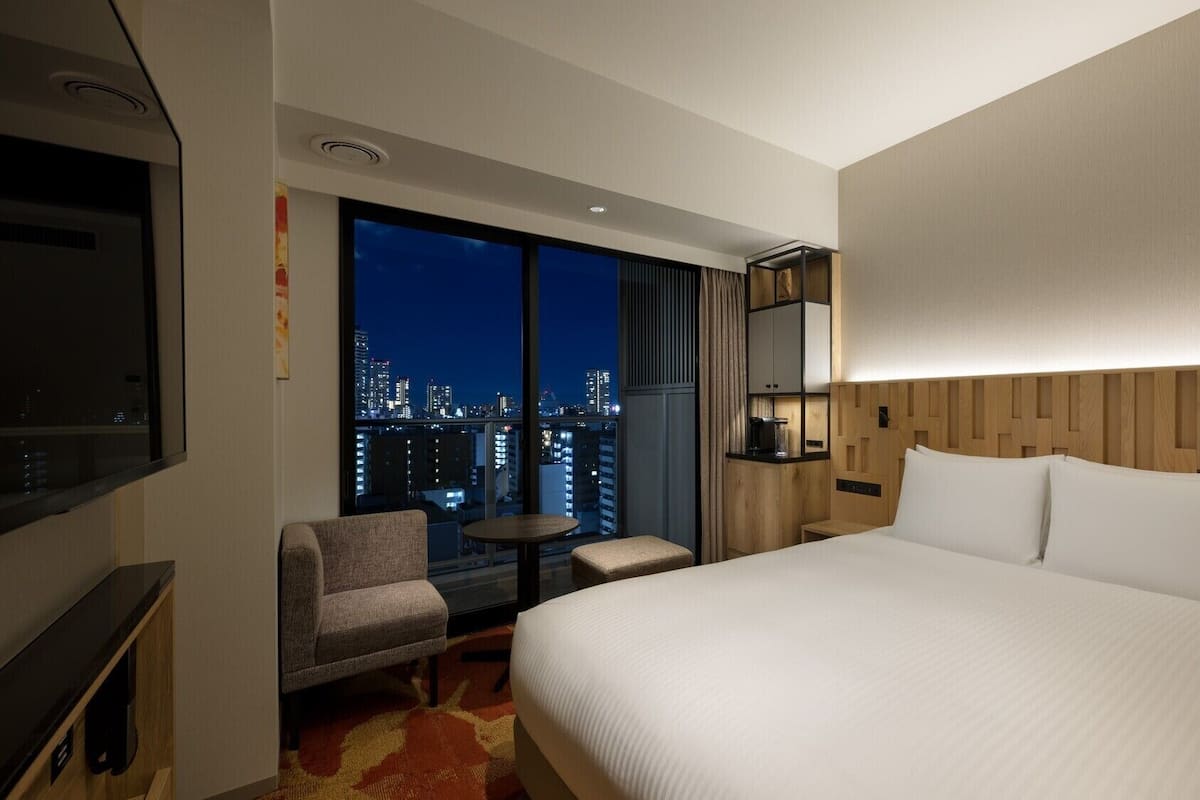 Hotelkamer met een groot bed, een televisie, een tafeltje, een stoel en uitzicht op de stad 's avonds.