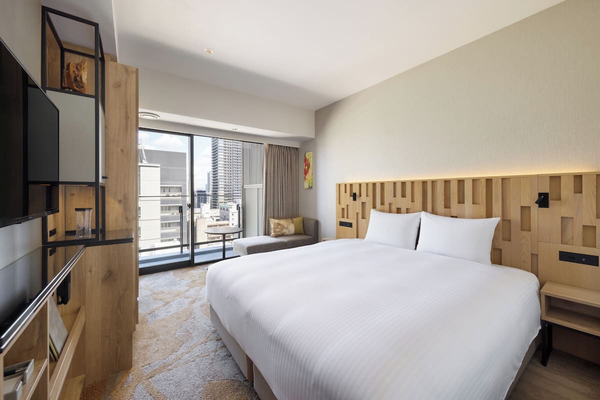 Een moderne hotelkamer met een groot bed, een houten hoofdeinde, een televisie en uitzicht op de stad door grote ramen.