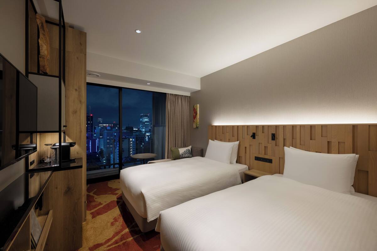 Een hotelkamer met twee bedden, een televisie, een bureau met lamp en een groot raam met uitzicht op de stad.