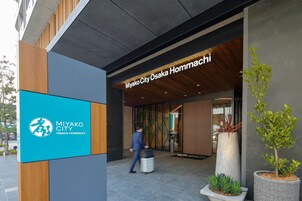 Der Eingang des Hotels Miyako City Osaka Hommachi mit moderner Fassade, einem Schild und Topfpflanzen.