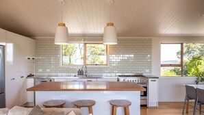 Una cocina moderna con armarios blancos, una isla de madera con taburetes, iluminación colgante y amplias ventanas que ofrecen vistas al verdor.