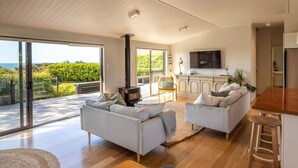 Amplia sala de estar con un gran ventanal corredizo que da a un balcón, chimenea y zona de cocina con televisor.