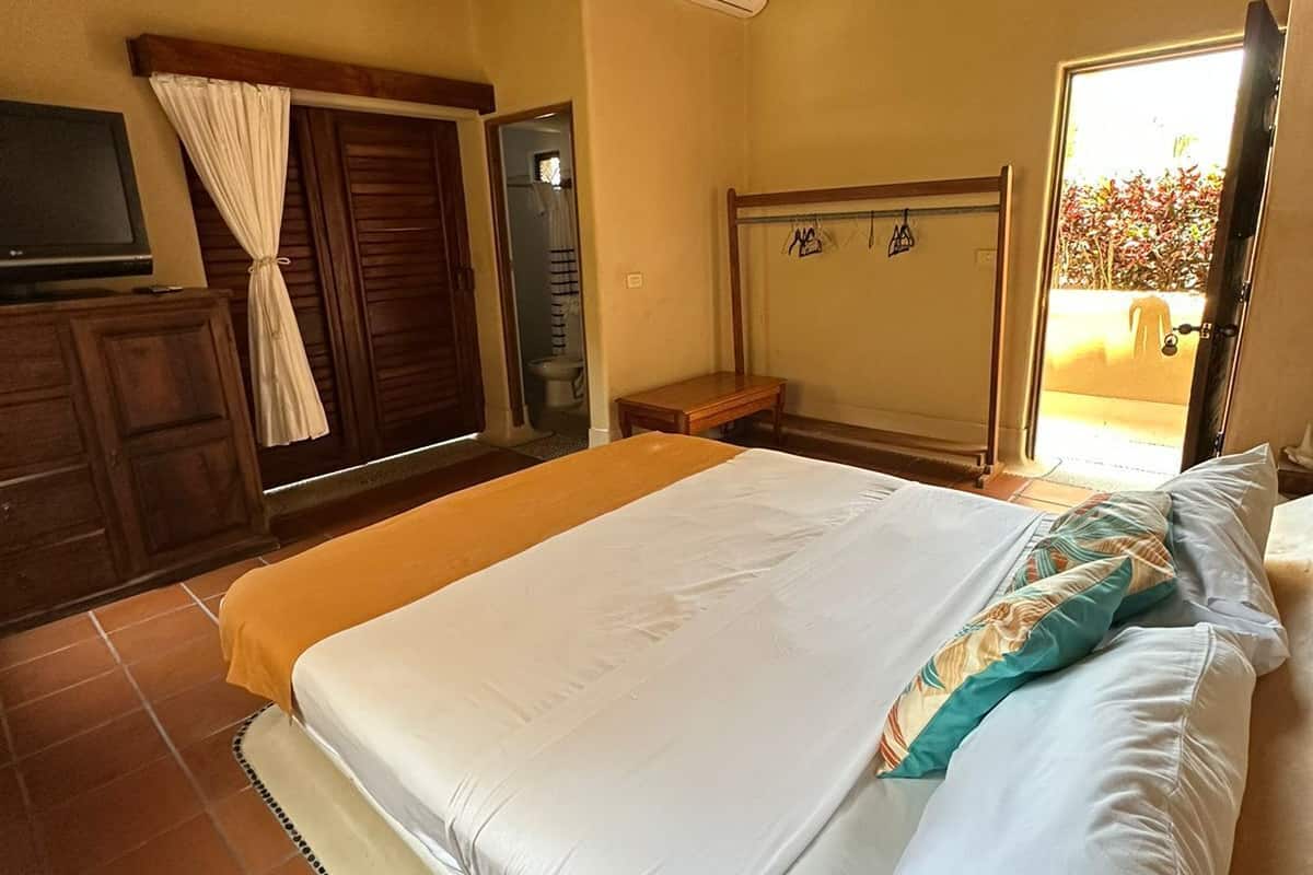 Un dormitorio con cama, ventilador de techo, televisión y ventana con vista a follaje.