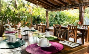 An outdoor dining area with wooden tables and chairs, surrounded by lush greenery.