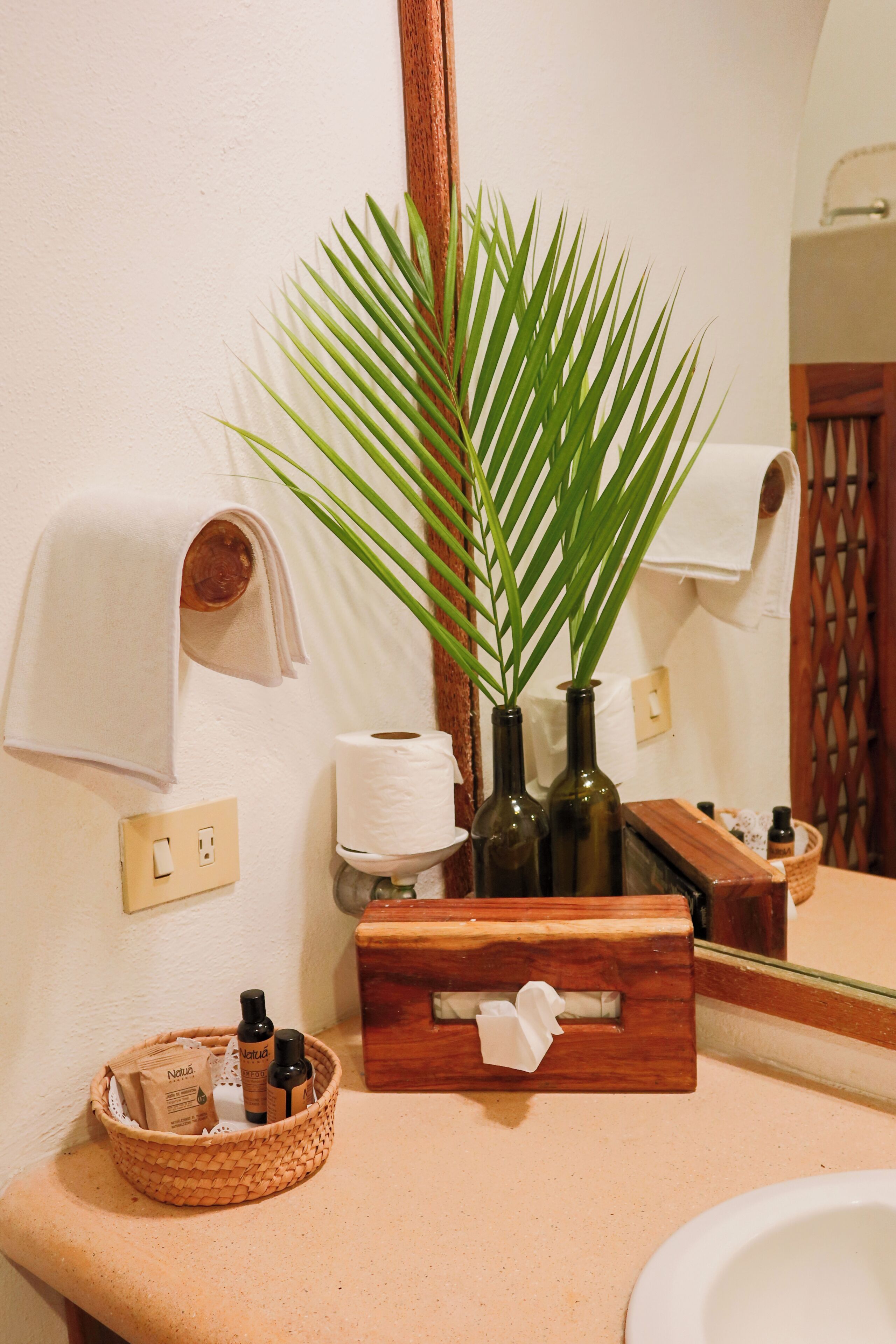 Een badkamer met een houten wastafelmeubel, een spiegel, een wastafel, een handdoekhouder, een plant en een mandje met toiletartikelen.