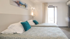 A hotel room with two beds, each with a patterned bedspread and teal pillows. There is a painting on the wall, a window with curtains, and an air conditioning unit. - 2 bedroom nice apartment in Algajola (Algajola)